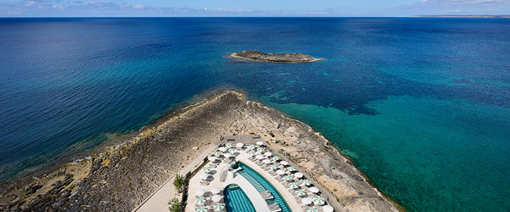Drohnenaufnahme der Poolanlage im Iberostar Selection Es Trenc mit Blick auf das türkisblaue Meer und eine kleine Insel.