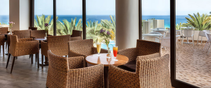 Gemütlicher Hotel-Loungebereich mit Korbsesseln hinter großen Panoramafenstern, dekoriert mit Cocktails und Blick auf die tropische Gartenanlage.
